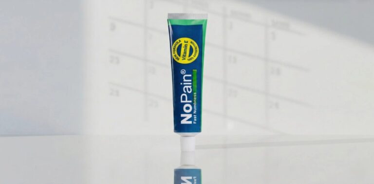 A centered, upright close-up shot of a white tube of "NoPain Cream" for "Relief & Recovery" with a multi-colored gradient bar. It stands on a highly polished white surface, creating a clear mirror image of the tube. The background is a blurred white wall with faint grid-pattern numbers (resembling a calendar) and complex, dappled light-break shadow patterns.