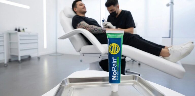 A sharp foreground product shot of a tube of 'NoPain®' numbing cream in a metal tray. The blue and green tube has large white vertical text: 'NoPain®' and 'Fast Numbness', plus a circular badge stating 'NEW FORMULA, Vitamin E, 100% SATISFACTION'. In the softly focused background, a man with intricate arm tattoos is reclining in a white tattoo chair, getting tattooed by an artist in black in a clean, modern studio.
