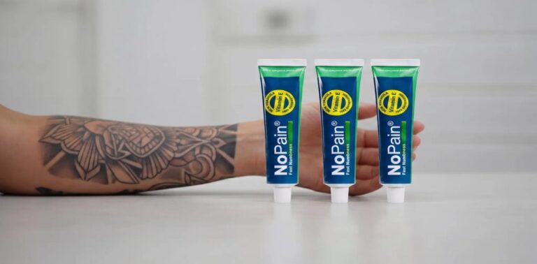 Three tubes of NoPain numbing cream standing in a row on a light surface. To the left is a tattooed forearm, and behind the tubes, a hand is visible against a simple, blurred white background