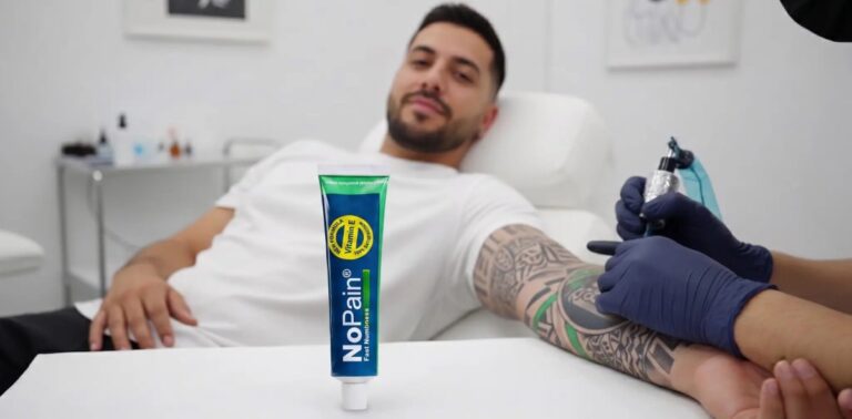 A tube of NoPain numbing cream standing on a white table, with a man getting a tattoo on his arm by an artist in the background